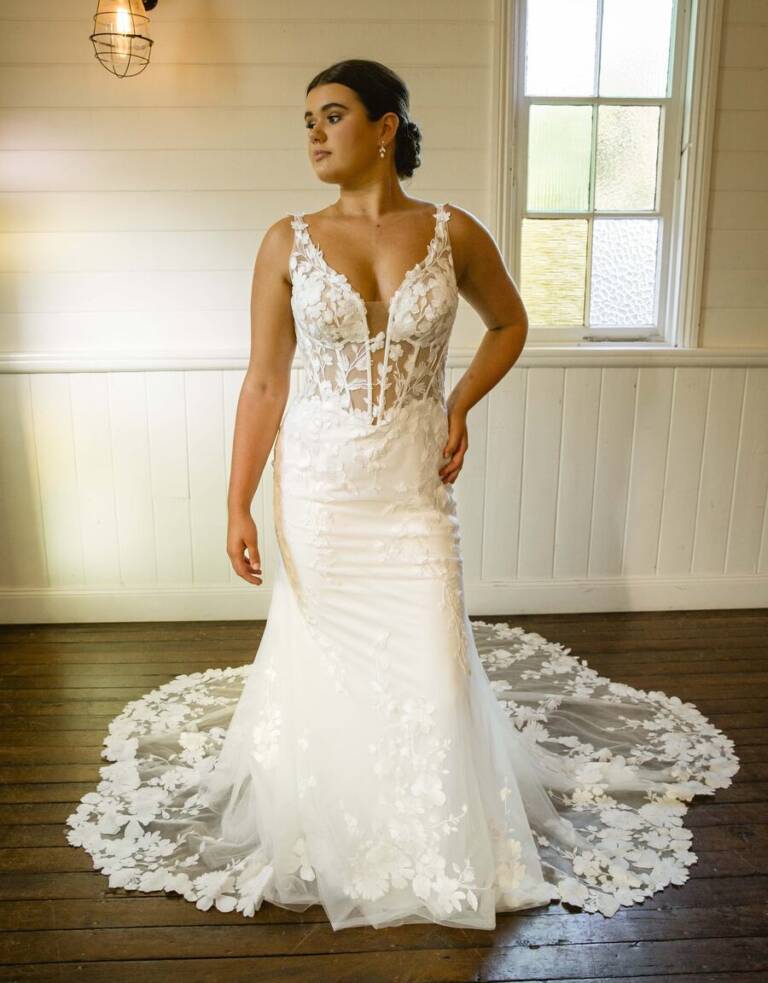 Bride wearing the Caris wedding gown from the RSVP Bridal Collection, featuring a sheer floral lace bodice, plunging neckline, and a dramatic lace train, standing in a softly lit room with vintage decor.