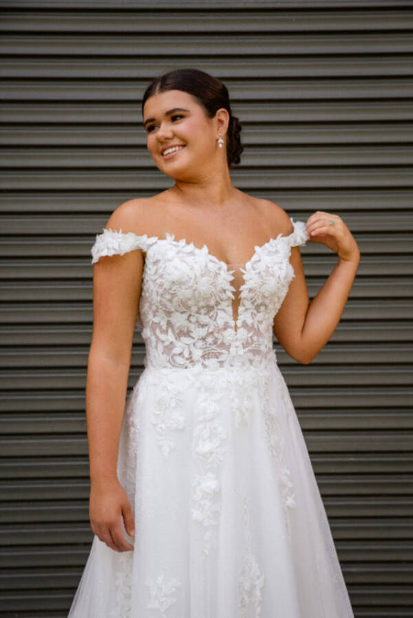 Bride wearing the Spring wedding dress from the RSVP Bridal Collection, featuring off-the-shoulder lace straps, a floral appliqué bodice, and a flowing A-line skirt with a chapel-length train.