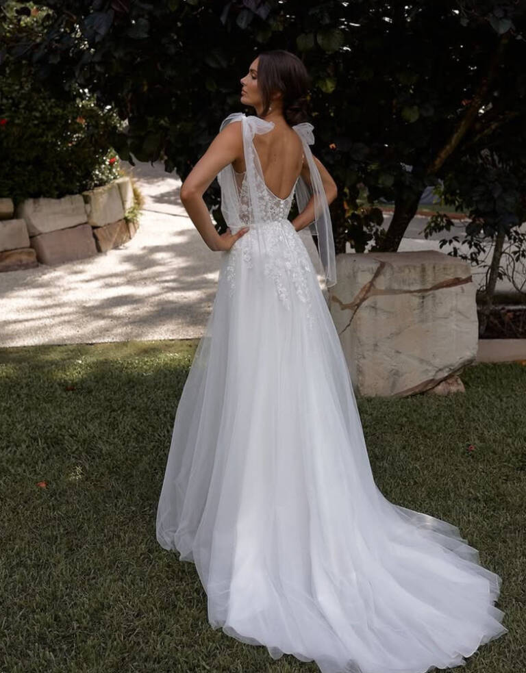 Ethereal beaded lace and tulle wedding dress in ivory with low V sheer back, tulle bows on shoulders, beaded lace applique cascading over waist. Soft tulle skirt flowing to a short train. Back view