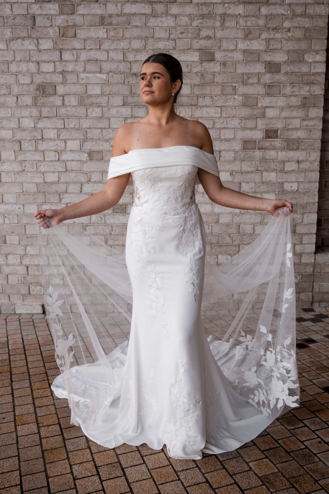 Bride wearing the Aria wedding dress, featuring an off-the-shoulder neckline and floral embroidery with sheer tulle wings, standing against a neutral brick wall.