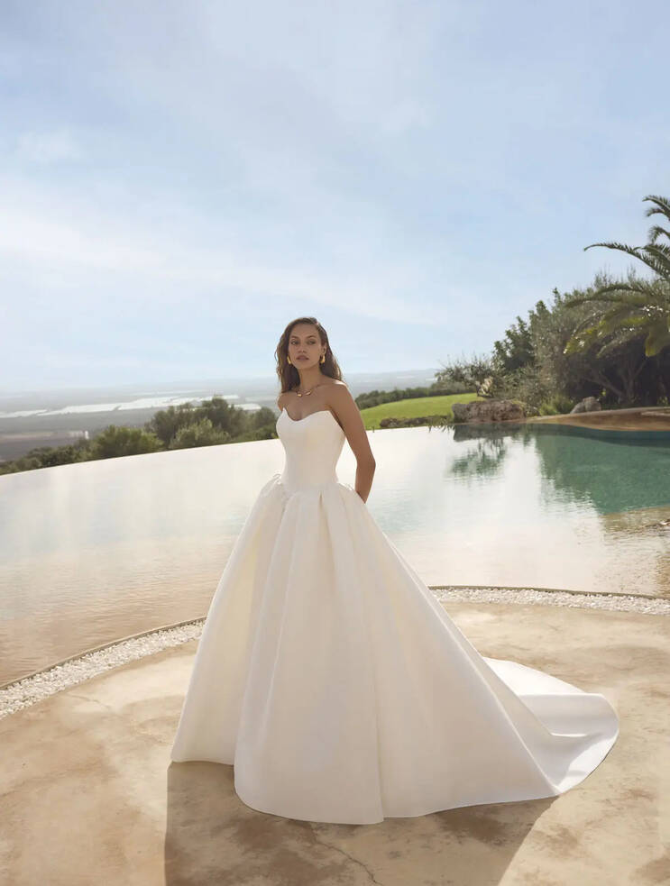 Bride wearing the Ghada mikado strapless ballgown by Modeca with a full skirt and scoop neckline, standing beside a pool under a clear sky.