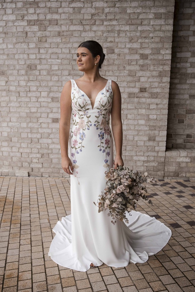 Bride wearing the RSVP Dannika ivory crepe fit-and-flare wedding dress with pastel floral lace panels and V-neckline, standing against a light brick wall holding a dried bouquet.