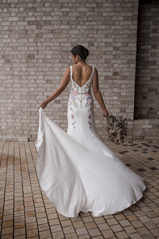 Back view of the RSVP Dannika ivory crepe wedding gown showing the low V-back, trailing pastel floral lace and flowing train as the bride lifts the skirt slightly.