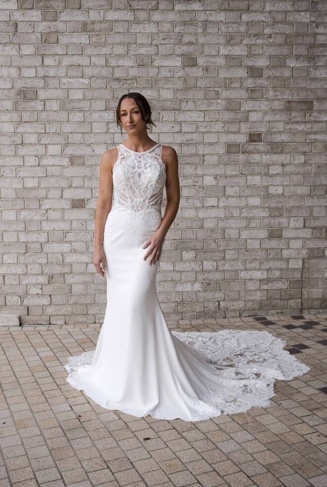 Bride wearing the RSVP Dana ivory crepe fit-and-flare wedding dress with sheer beaded lace bodice and illusion neckline, standing against a light brick wall.
