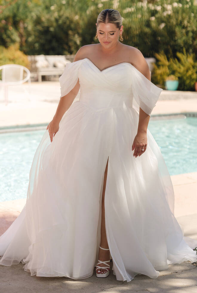 Front view of plus-size bride in Stella York Style SY8215+ off-the-shoulder ivory organza wedding gown with sweetheart neckline, basque waist and thigh-high leg split, standing by a pool.