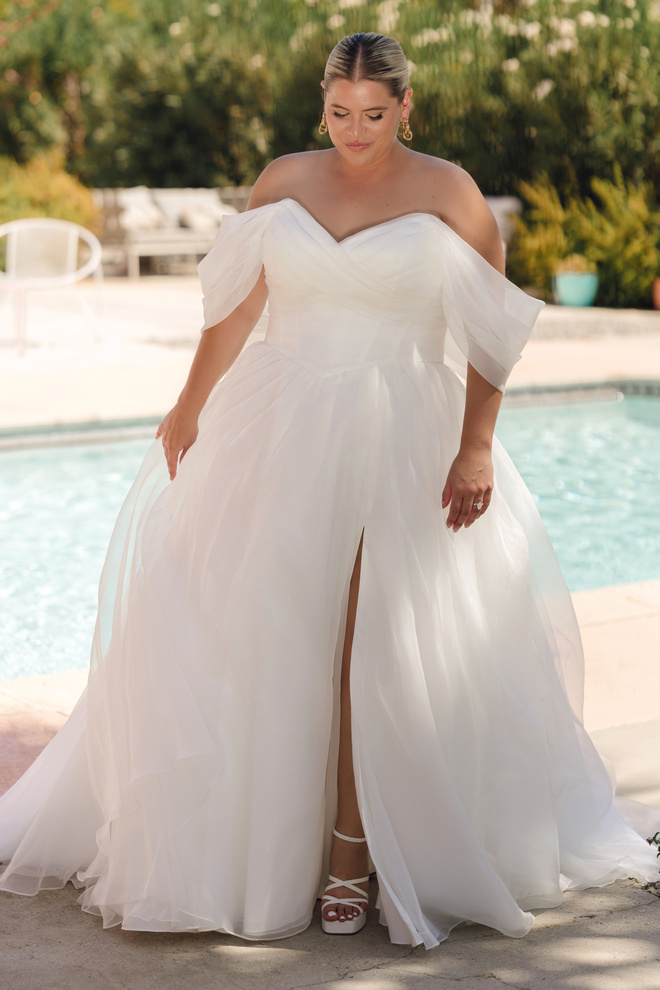 Front view of plus-size bride in Stella York Style SY8215+ off-the-shoulder ivory organza wedding gown with sweetheart neckline, basque waist and thigh-high leg split, standing by a pool.