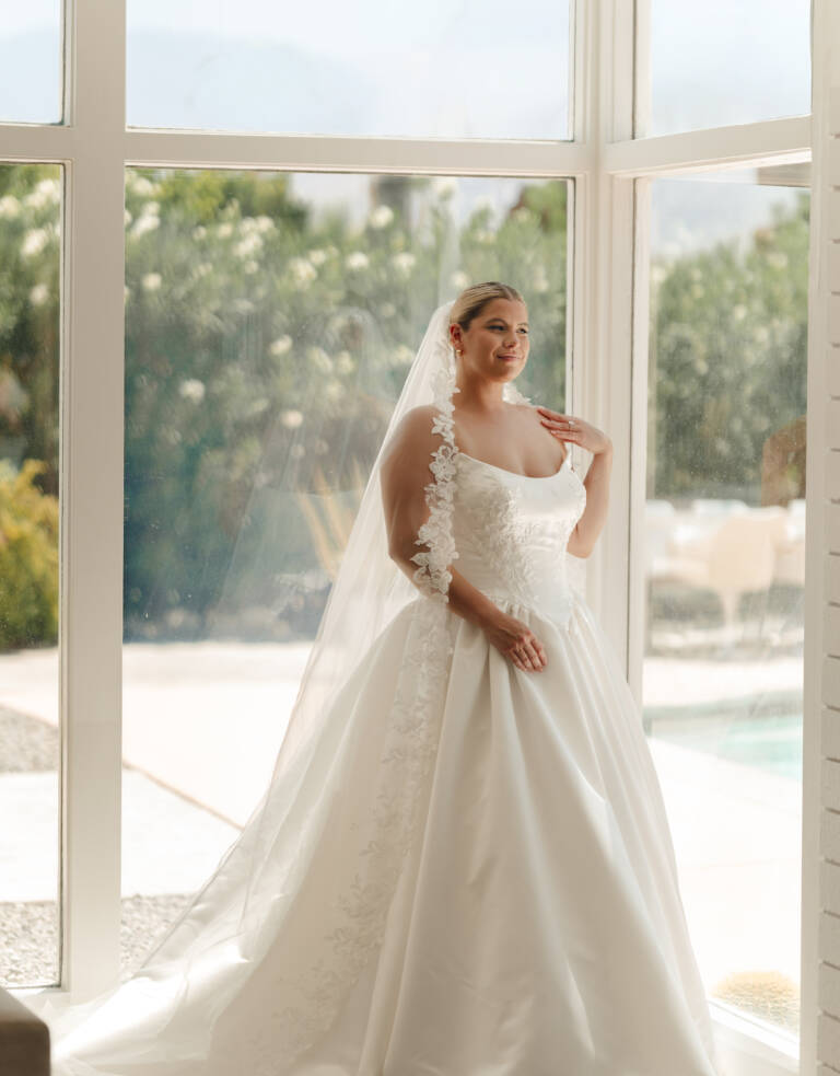 Plus-size bride wearing Stella York SY8239+ strapless satin ballgown, standing by sunlit floor-to-ceiling windows showing cat-eye neckline and full skirt.