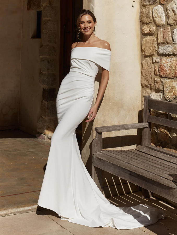 Model wearing the Abbie TC2639 wedding dress by Tania Olsen Designs — a white stretch crepe fit-and-flare gown with a straight neckline, soft pleating from the left hip, and an elegant off-shoulder look.