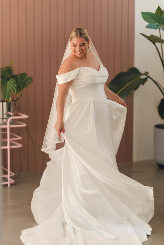 Bride wearing Stella York SY8236+ satin A-line wedding dress with off-shoulder pearl neckline, Basque waist and flowing satin skirt.