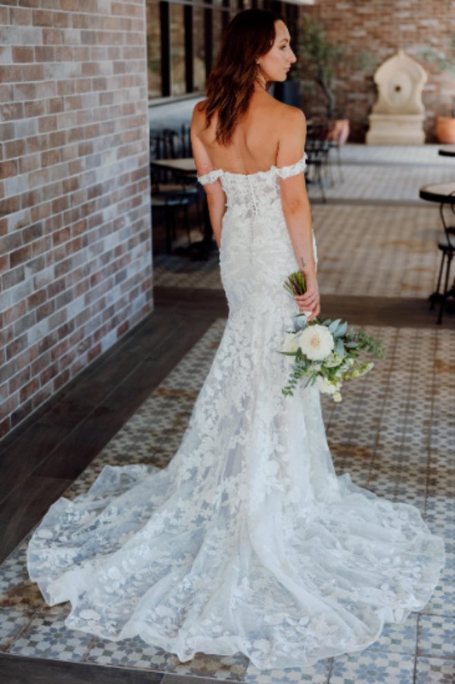 Back view of the Heidi wedding dress showcasing off-the-shoulder straps, intricate lace detailing, fabric-covered buttons and a flared lace train.